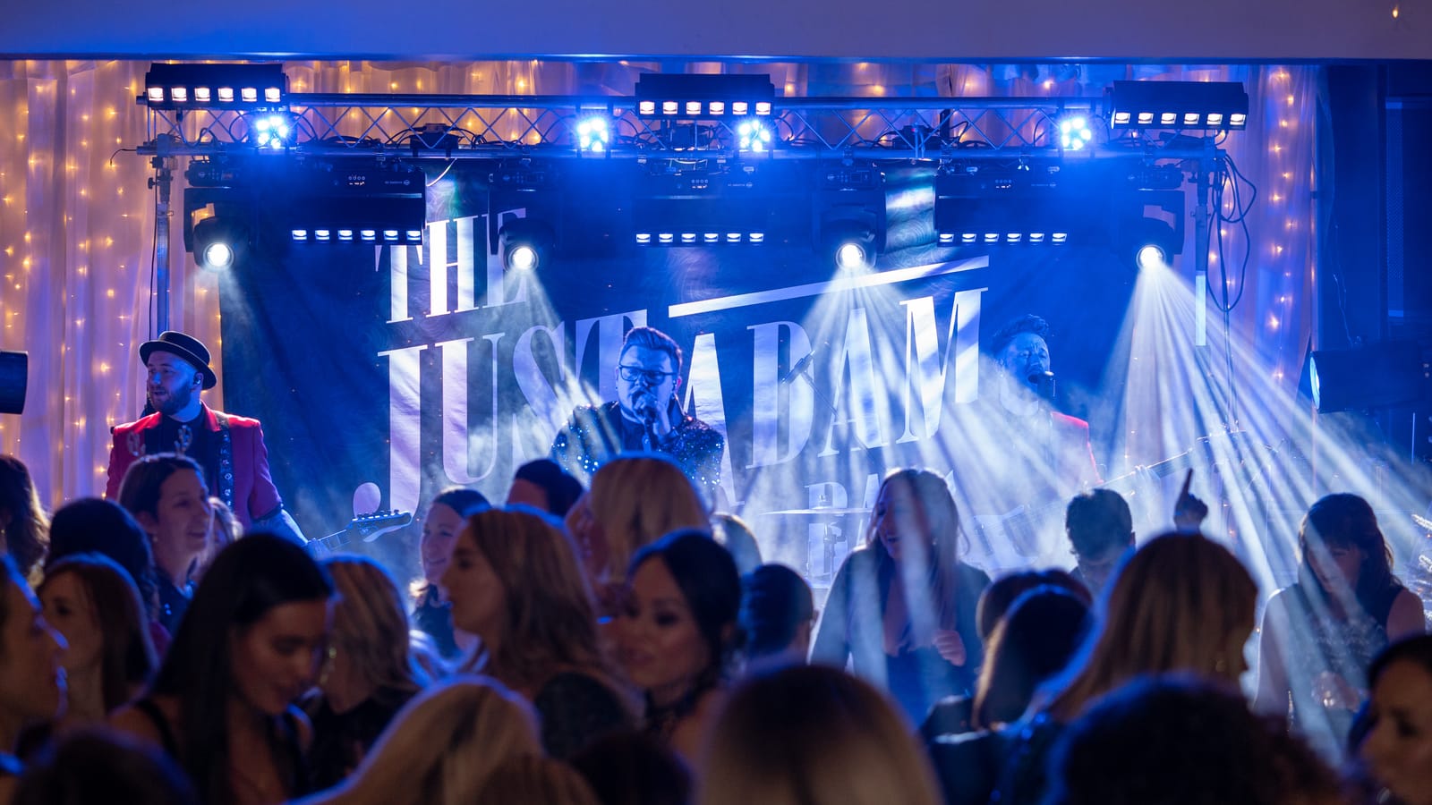 A full dance floor in front of the band, lit blue, under The Just Adam Band backdrop